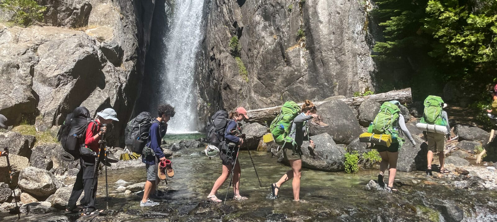 Youth Backpackers cross a stream in front of a beautiful waterfall.