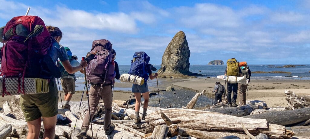 Teens backpacking over driftwood with sea stacks in the background in Olympic National Park