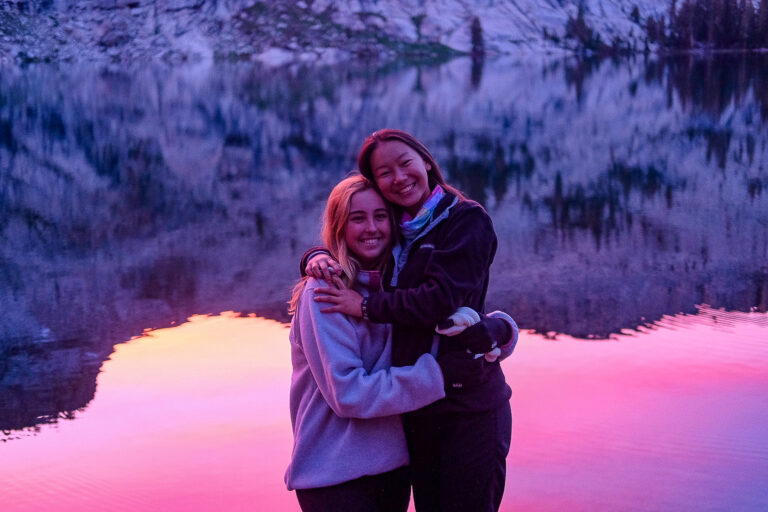 Girls hugging in sunset reflection on lake