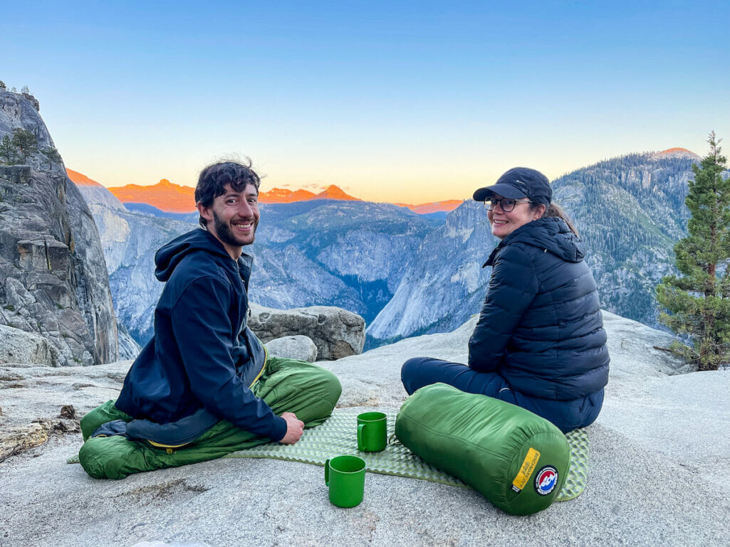 Couple shares a moment watching the sunset with sleeping bags and hot beverages.