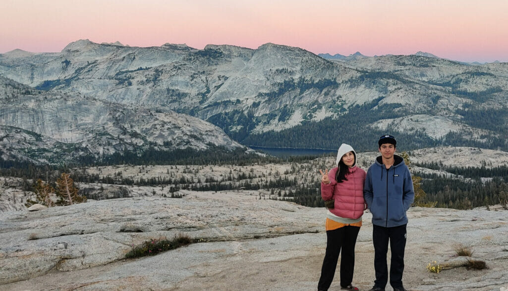 A mother and son pose for the camera in front of a pink sunset on the bluff at May Lake.