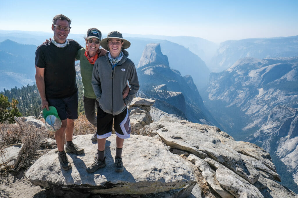 A family smiles on the summit of Clouds Rest with Half Dome in the background.