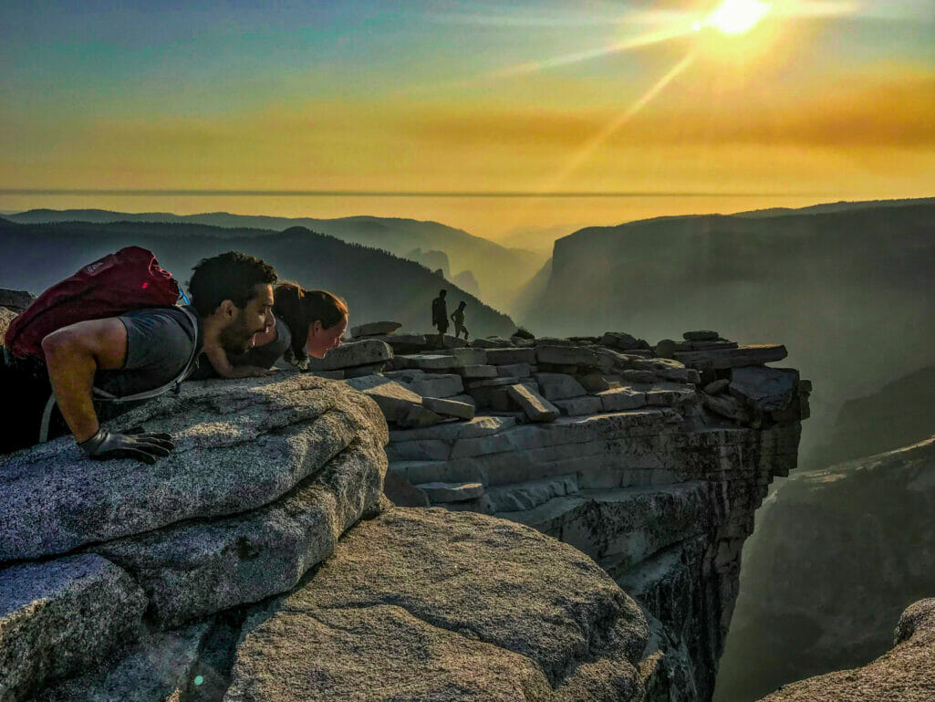 Two people peer over the edge of Half Dome as two others walk to the edge.