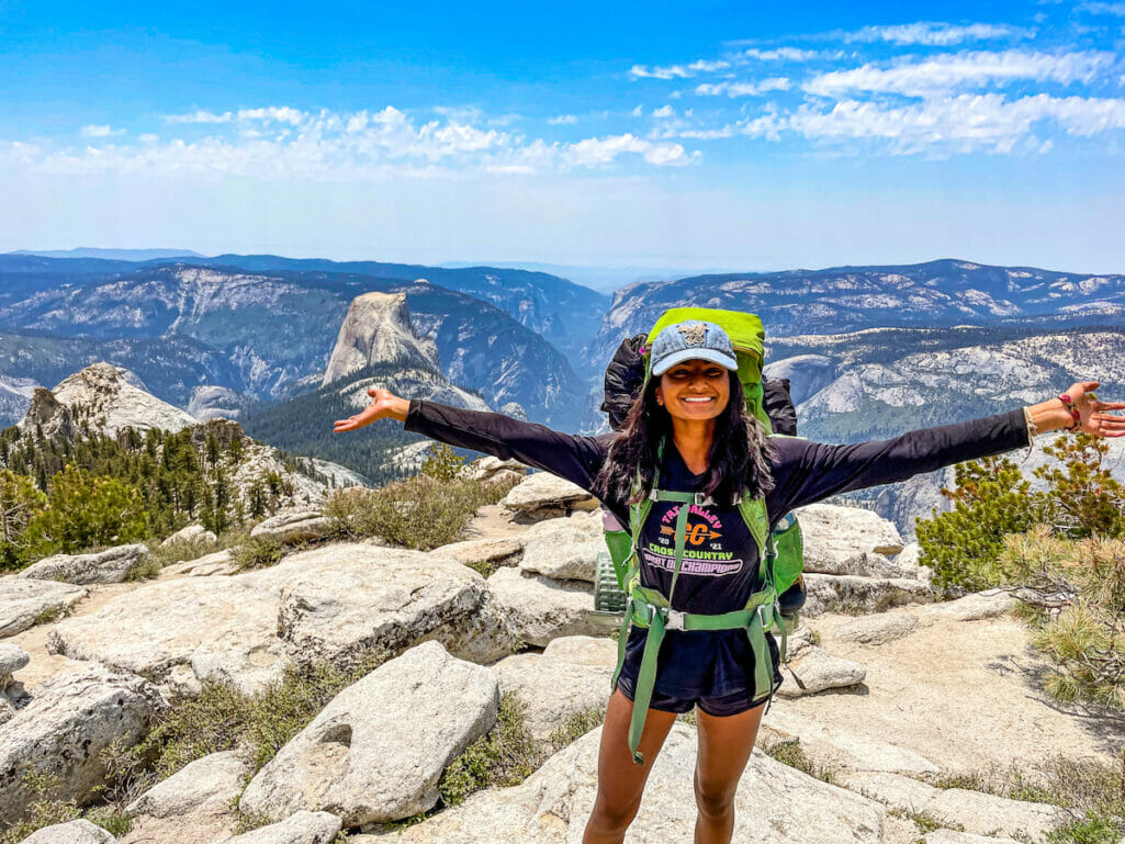 A smiling backpacker on top of Clouds Rest with Half Dome in the background.