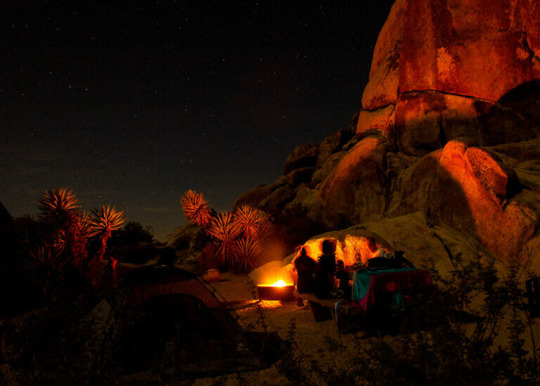 camping under the stars and around a campfire at Joshua Tree National Park. The boulders and joshua trees glow red from the fire.