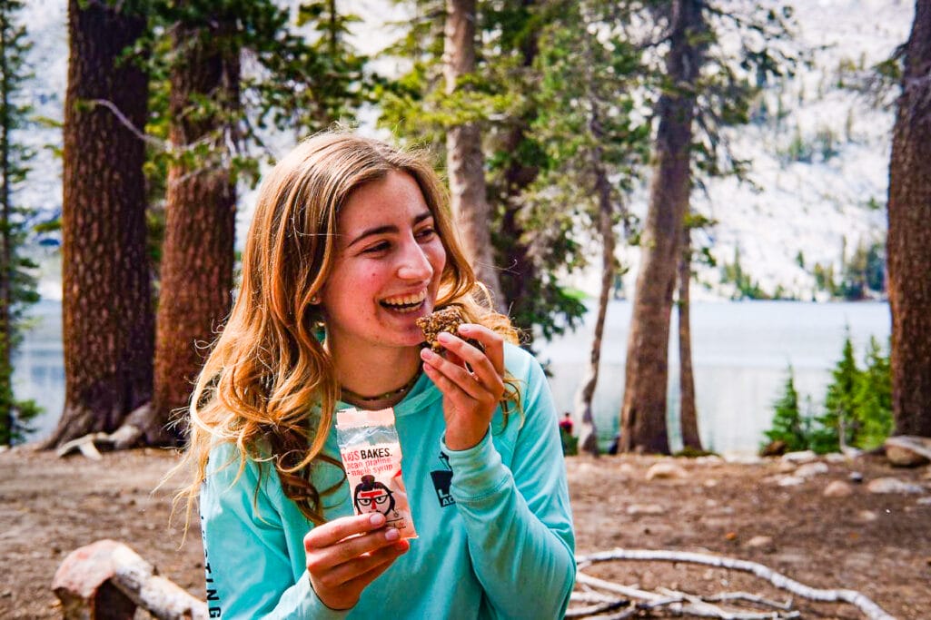 Woman eats a Taos Bakes bar in front of a lake in Yosemite National Park.